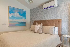 Ein Schlafzimmer mit einem großen Bett, einem Kopfteil, einer wandmontierten Klimaanlage und einem Gemälde mit Strandmotiv.