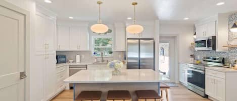 Una cocina moderna con una isla central, taburetes de bar, electrodomésticos de acero inoxidable e iluminación colgante.