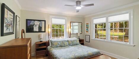 Un dormitorio con cama, mesitas de noche, un escritorio y una silla. Se ven vistas de vegetación por las ventanas.