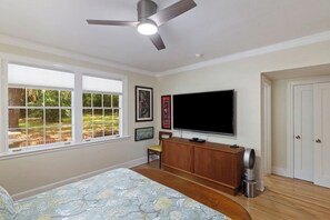 Ein Schlafzimmer mit einem Bett, einem Fernseher, einem Deckenventilator und einem Fenster mit Blick auf Grünflächen.