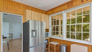 A compact kitchen with wooden paneling, a stainless steel refrigerator, a microwave, and a window with a view of greenery. - Quaint and Quiet near UF! (Gainesville)