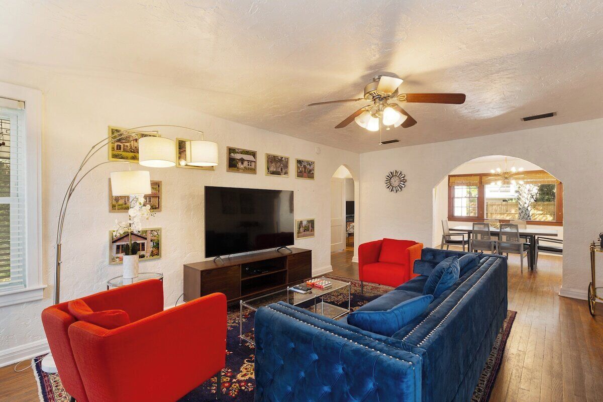 A living room with a television, red and blue sofas, a ceiling fan, and a dining area with a table and chairs.