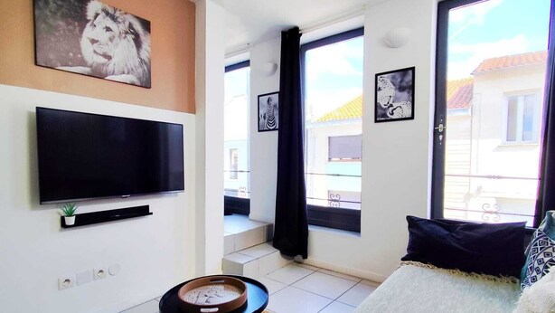En stue med et fladskærms-tv, et lille bord, en sofa og et vindue med udsigt til bygninger.