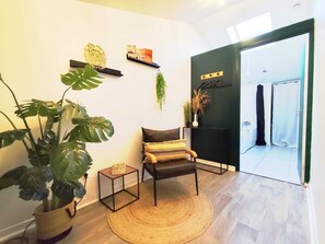 Ein Zimmer mit Holzboden, einem runden gewebten Teppich, einem Sessel mit gemustertem Kissen, einer Topfpflanze, einem kleinen quadratischen Tisch und einer Tür zu einem Badezimmer.