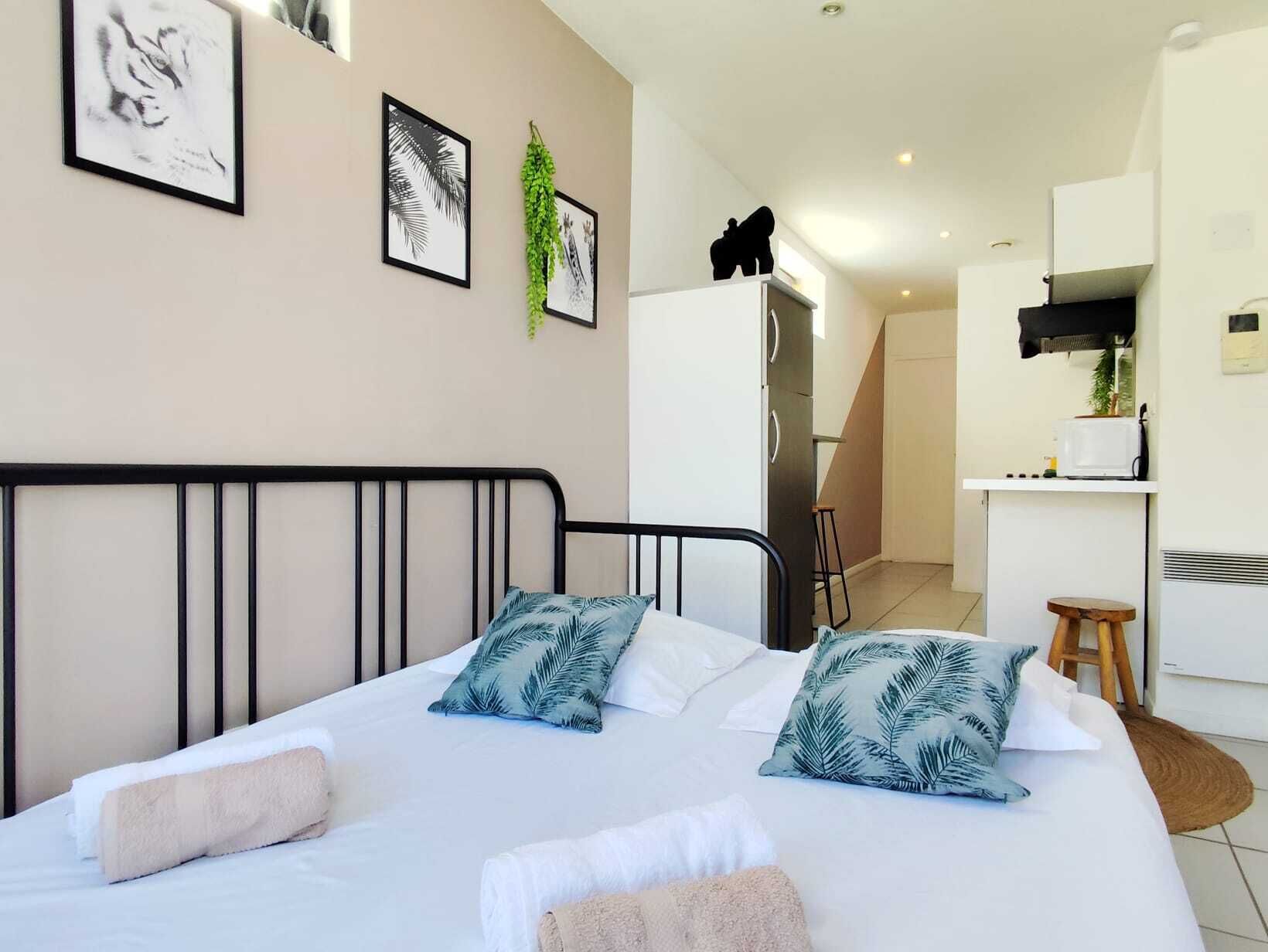 Une chambre à coucher avec un lit, des oreillers, une table de chevet, une plante et, au fond, une petite cuisine.