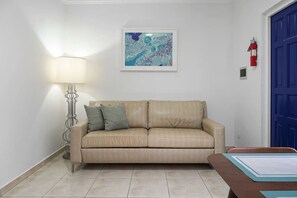 Ein beiges Ledersofa mit zwei Kissen, eine Stehlampe, ein gerahmtes abstraktes Gemälde, eine blaue Tür und ein Feuerlöscher an der Wand.