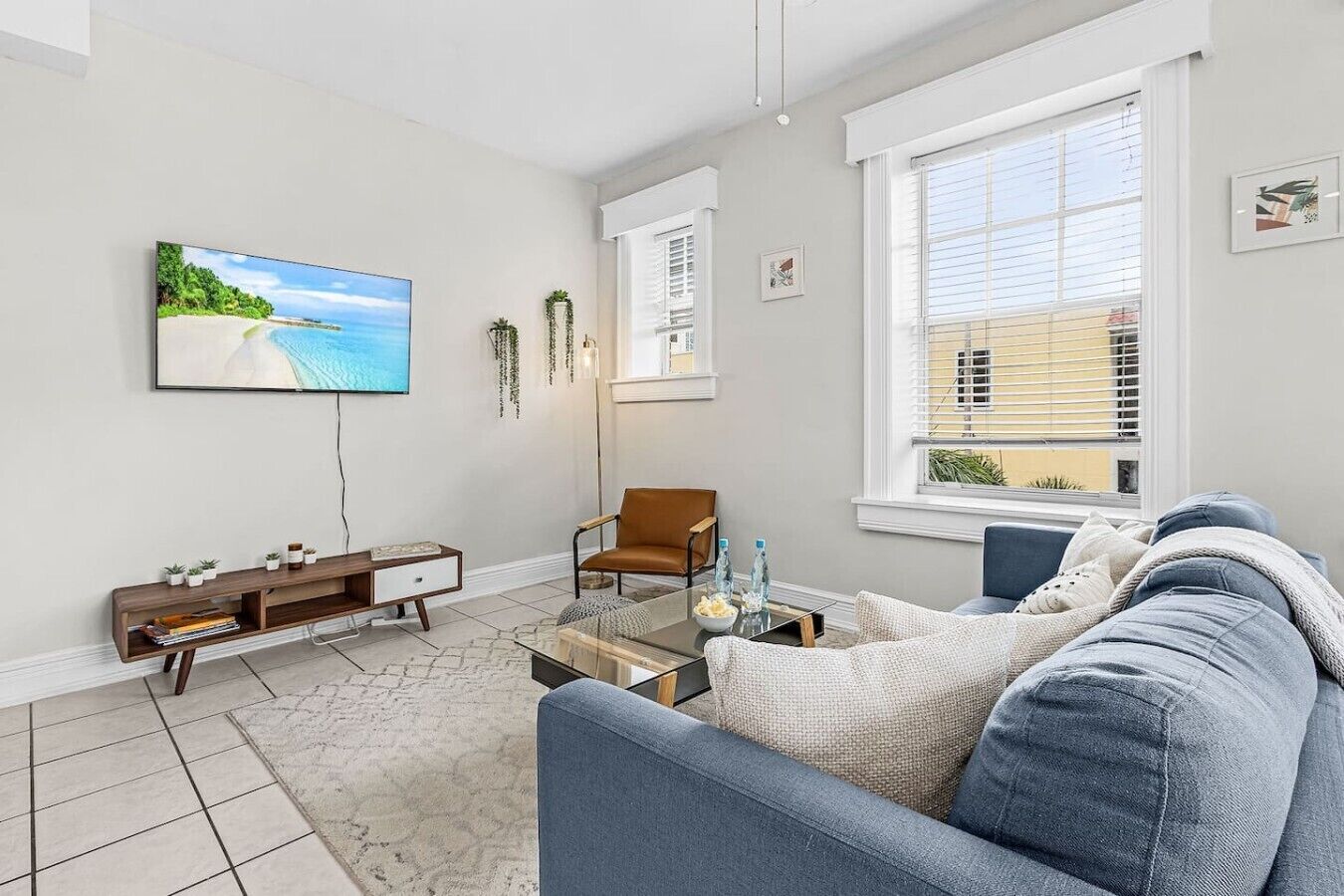 A modern living room with a sofa, a coffee table, a TV mounted on the wall, and a window with blinds.