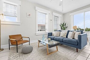 A living room with a blue sofa, a brown chair, a glass coffee table, and a large window with blinds.