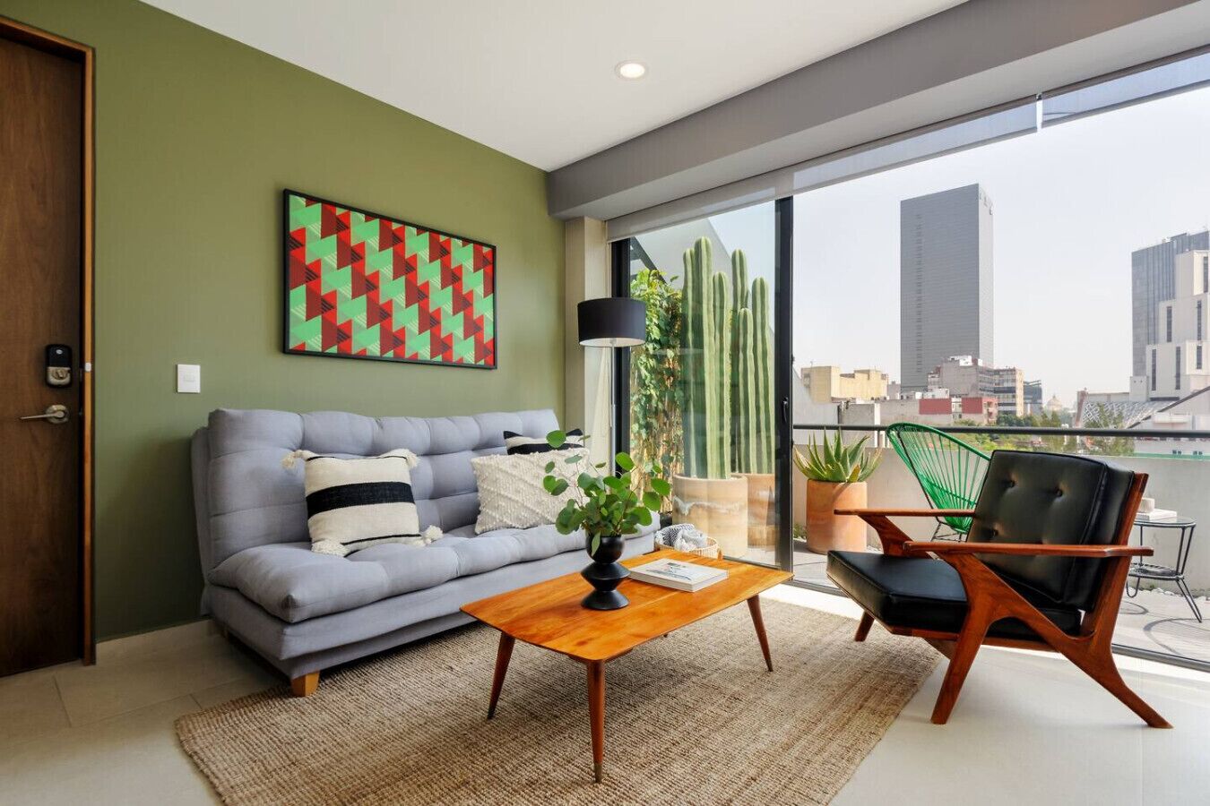 Ein modernes Wohnzimmer mit einem grauen Sofa, einem Holztisch und einem grünen Sessel. Ein großes Fenster bietet Blick auf Gebäude und eine Kaktusstaude draußen.