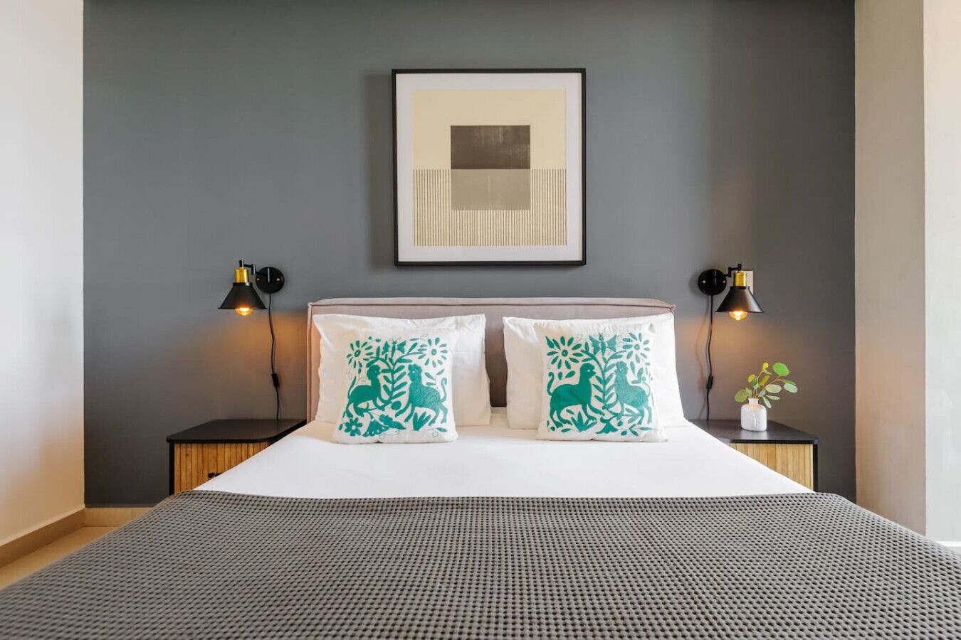 A neatly made bed with a grey textured bedspread, two pillows with green floral designs, and a framed abstract artwork above the bed.
