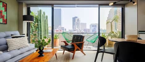 A modern living room with a sofa, coffee table, and chairs, a balcony with plants, and a city view.