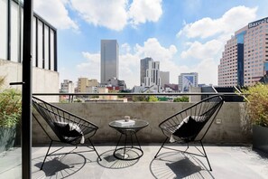 Terraço no telhado com duas cadeiras de estrutura em fio preto e uma pequena mesa redonda, com vista para uma paisagem urbana com diversos edifícios.
