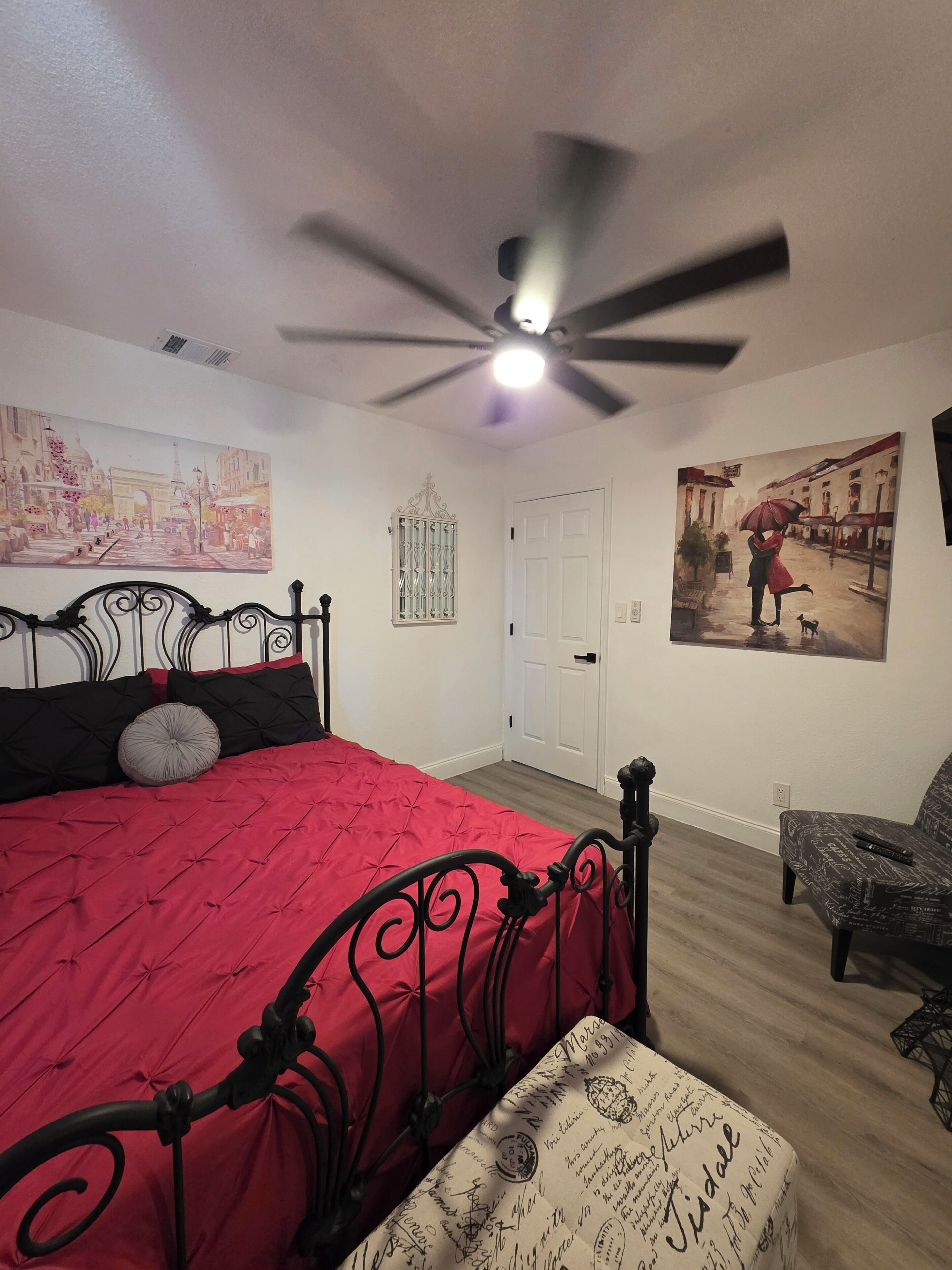 A bedroom with a bed, a ceiling fan, a door, and two wall-mounted paintings.