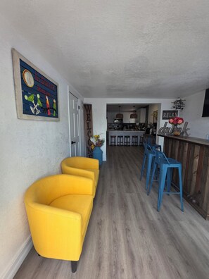 Ein Wohnzimmer mit gelbem Sofa, einer dahinterliegenden Küche und einer Bar mit Hockern.