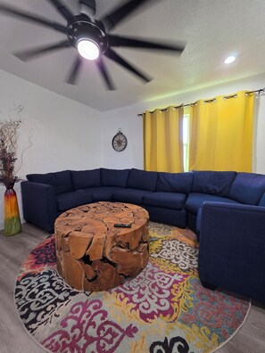 Ein Wohnzimmer mit einem blauen Moduls Sofa, einem Holztisch, einem bunten Teppich, einem Deckenventilator und einer Wanduhr.