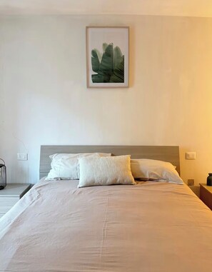 Cama bem arrumada com lençóis e almofadas brancas, uma planta emoldurada na parede e uma mesa de cabeceira com abajur.
