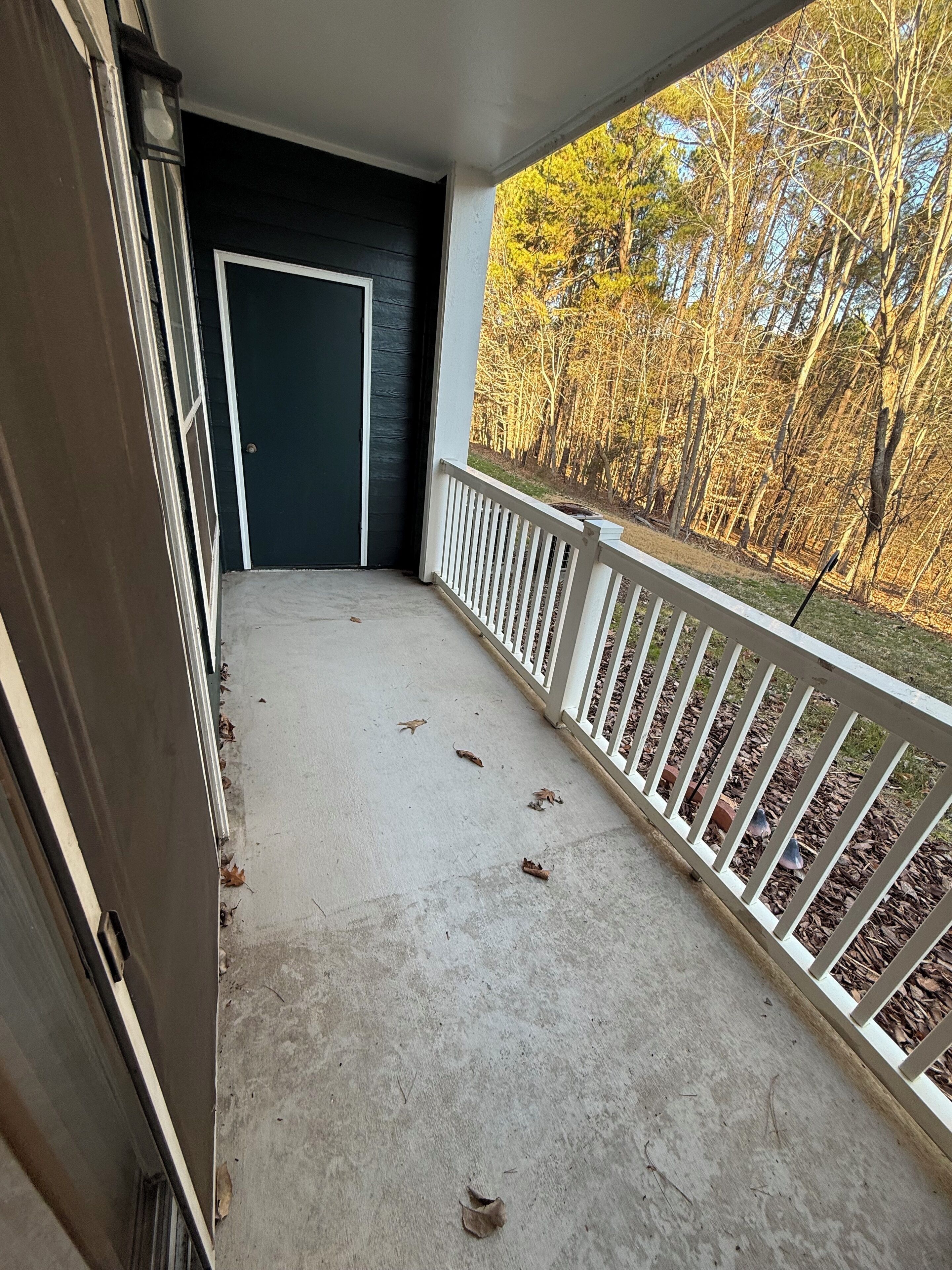 A covered balcony with a concrete floor, white railing, and a door leading to an interior space.
