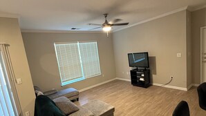 Ein Wohnzimmer mit Sofa, Deckenventilator, Fernseher und einem Fenster mit Jalousien.