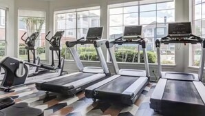 Ein Fitnessraum mit Laufbändern, Crosstrainern und Fahrrädern, großen Fenstern und einem strukturierten Boden.