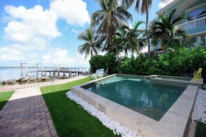 A swimming pool with a view of the sea, surrounded by palm trees and a building with balconies. - Luxury Bayfront Escape | Pool, Dock, Elevator & Sunset Views (Tavernier)