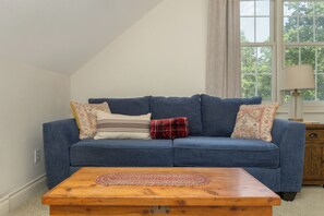 Ein blaues Sofa mit gemusterten Kissen, ein Holztisch und ein Fenster mit Blick auf Bäume.
