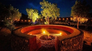 A circular stone fire pit with a central fire, surrounded by a low stone wall, under a night sky with trees and string lights.