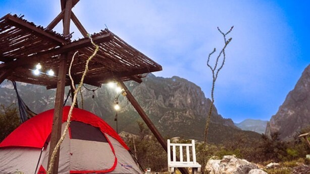 A tent with a wooden structure overhead, a white chair, and a rocky landscape with mountains in the background.