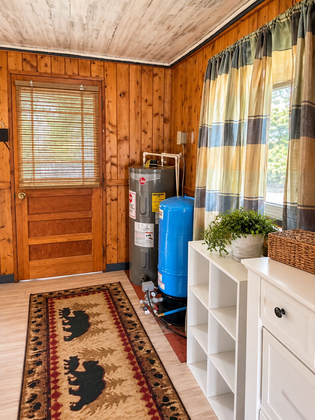 A room with wooden walls, a door, a window with blinds, a water heater, a blue water tank, a white cabinet, and a patterned rug.