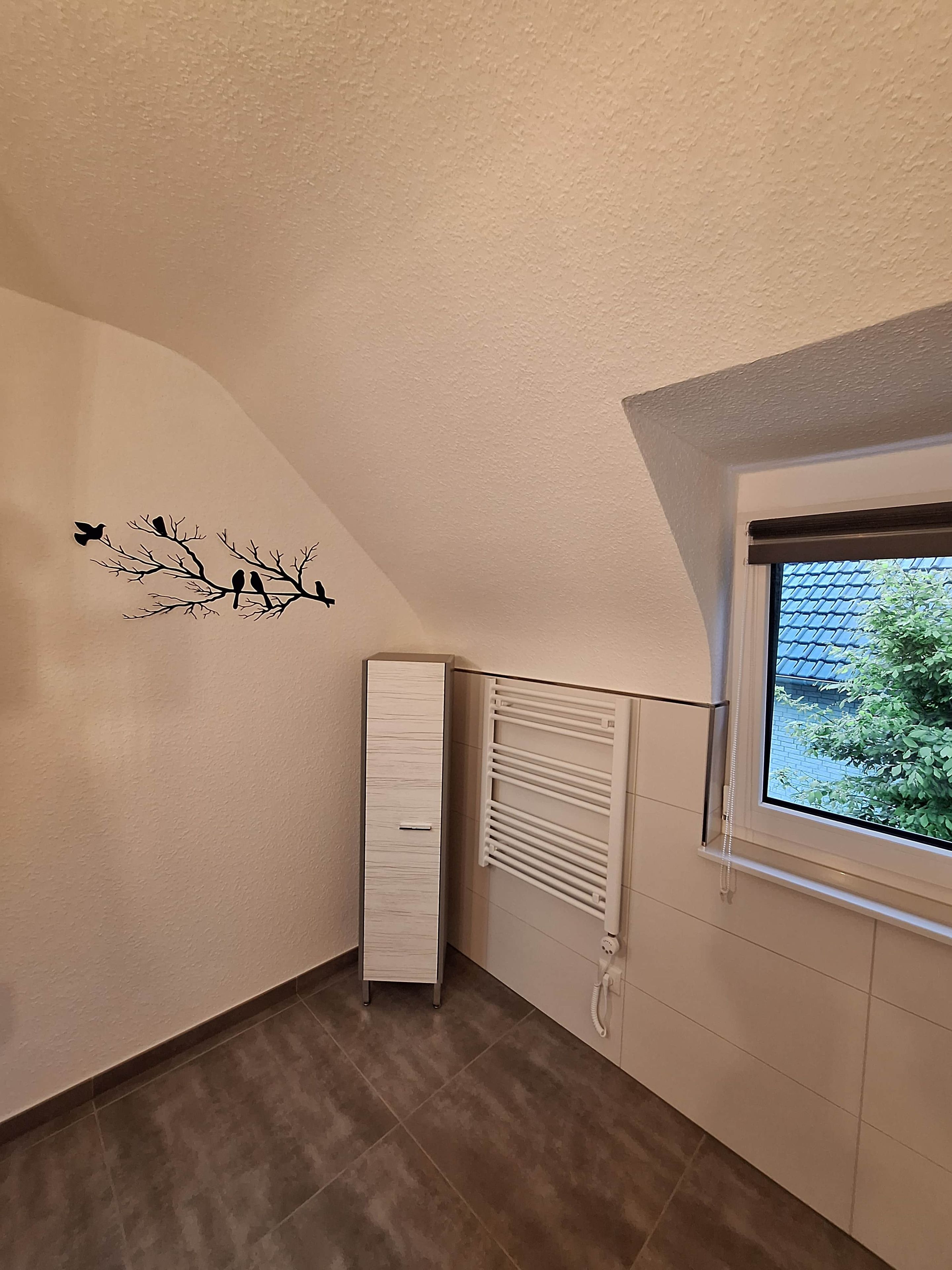 Una habitación con radiador, ventana con vista a vegetación y decoración de aves en una rama.