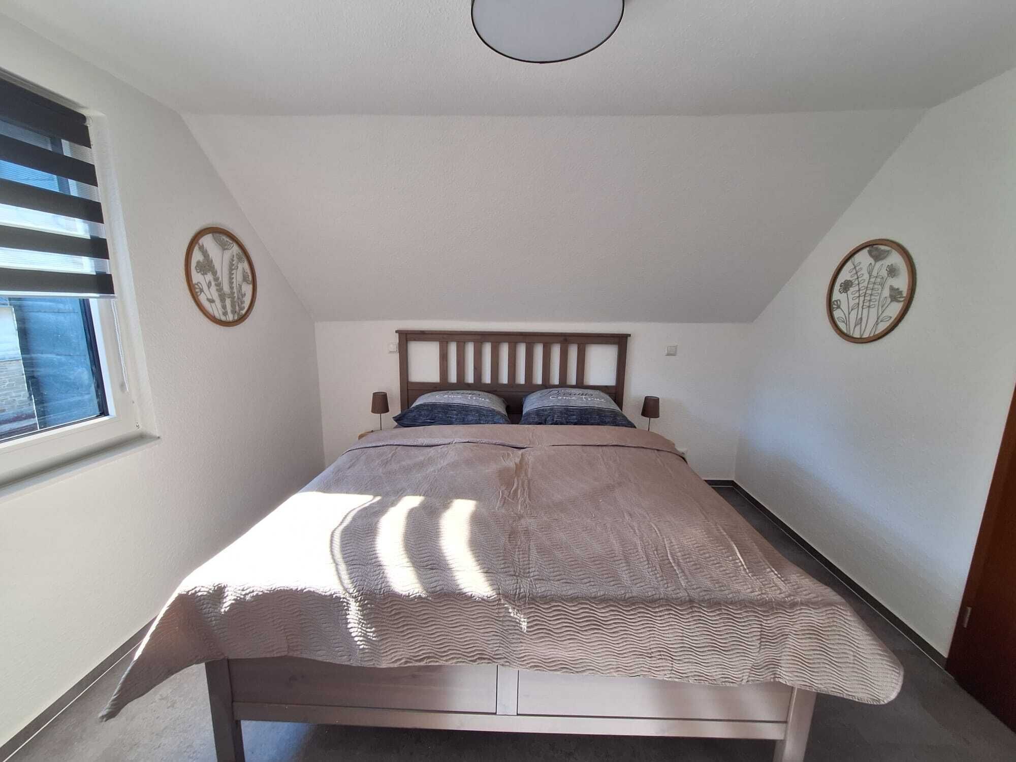 Un dormitorio con cama, dos mesitas de noche, una ventana con persianas y dos elementos decorativos en la pared.