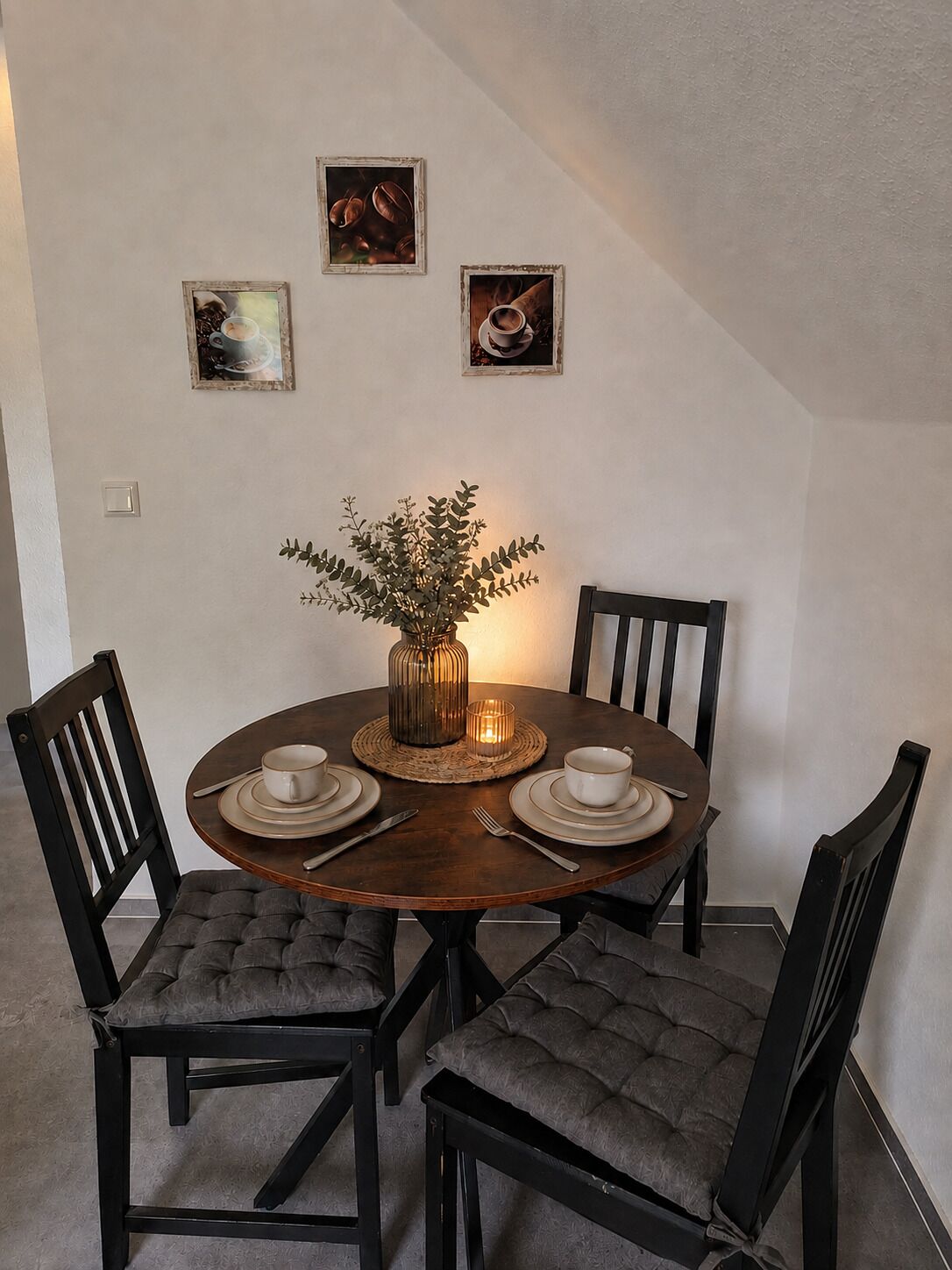 Un comedor con una mesa redonda, cuatro sillas, un jarrón con plantas y tres cuadros en la pared.