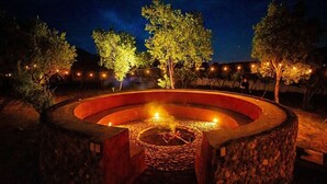 A circular stone fire pit with a central fire, surrounded by a low stone wall, under a night sky with trees and string lights.