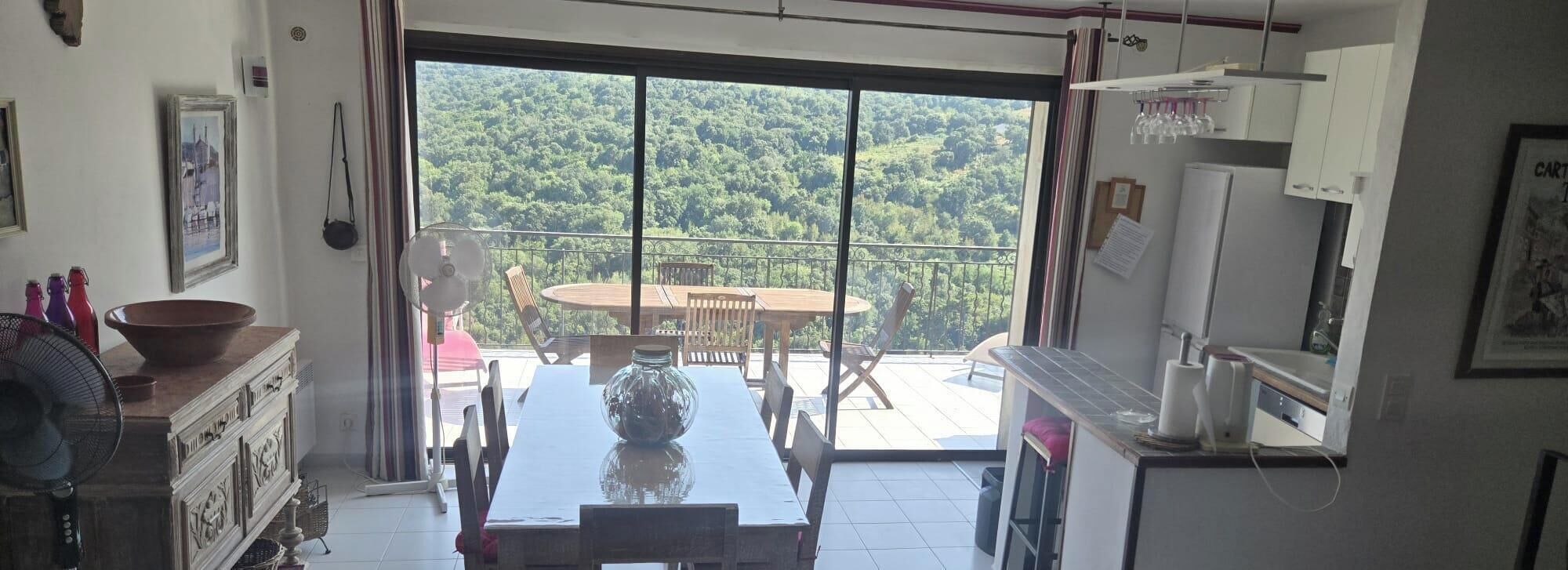 A room with a dining table and chairs, a large window with a view of a forest, and a balcony with a table and chairs.