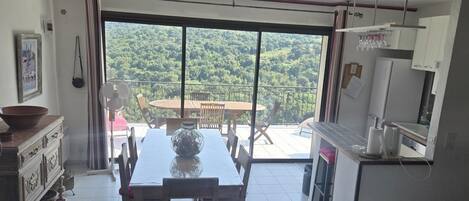 Une pièce comprenant une table à manger et des chaises, une grande fenêtre donnant sur une forêt et un balcon avec une table et des chaises.