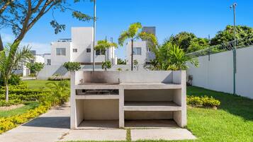 Un espacio exterior de hormigón con asientos, una banca y una mesa, rodeado de vegetación y un edificio blanco al fondo.