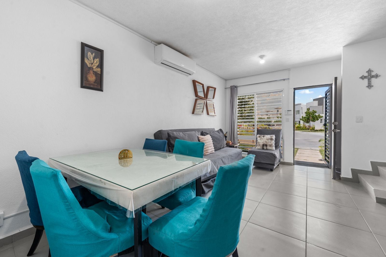 Una sala moderna con una mesa de comedor de vidrio, sillas color turquesa, un sofá gris y vista a una zona residencial a través de un ventanal amplio.