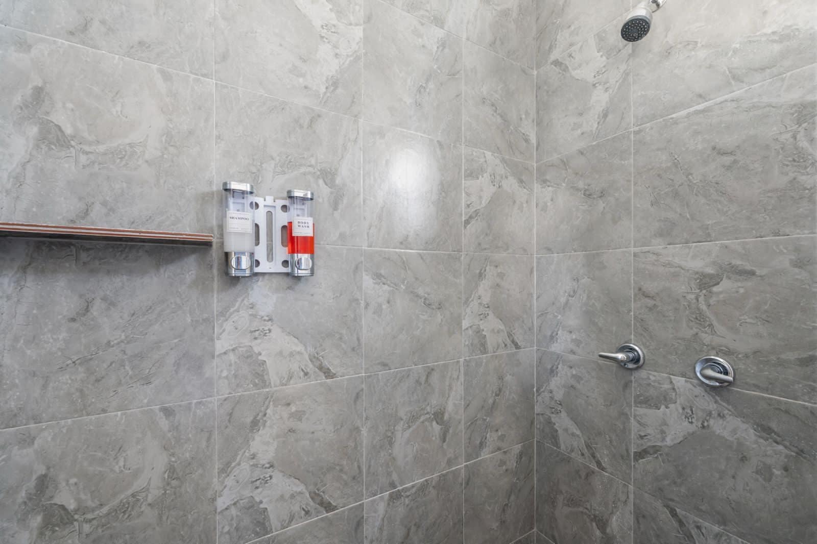Zona de ducha con azulejos de mármol gris, un cabezal de ducha y dos dispensadores fijos en la pared etiquetados 'champú' y 'gel de baño'.