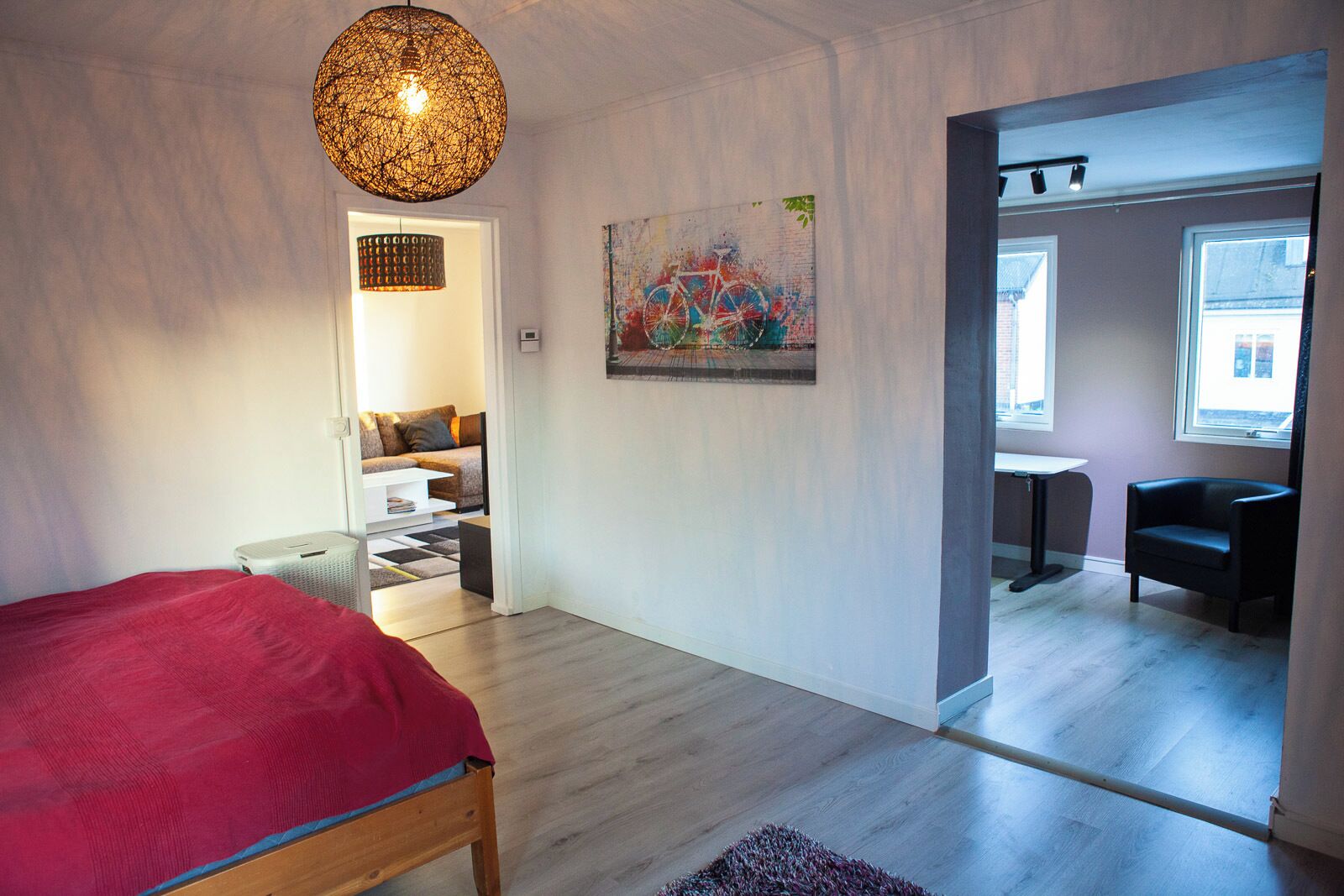 Ett modernt sovrum med en säng, en hängande lampa, en tavla och utsikt in i ett annat rum.