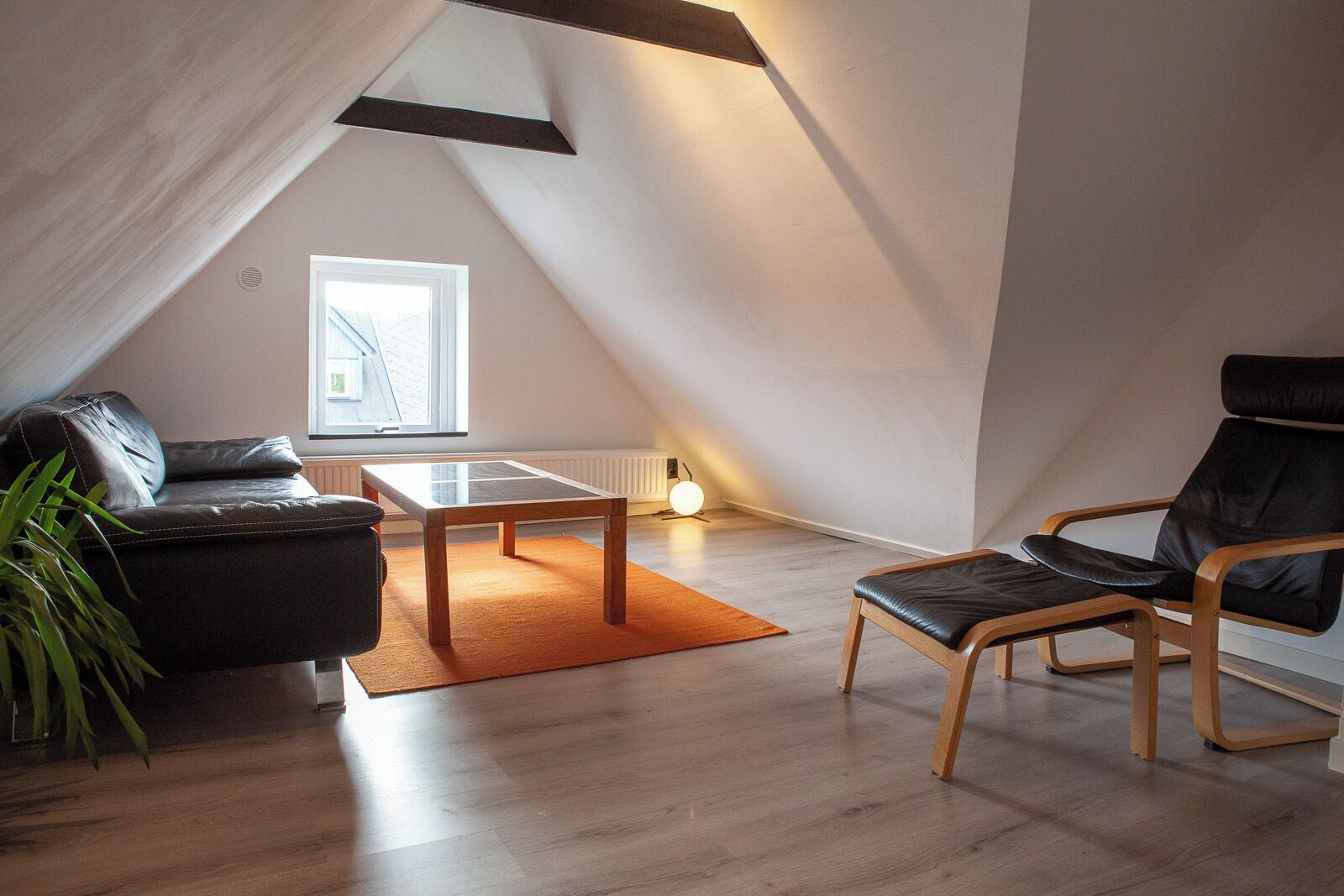 Ett modernt loftrum med en sluttande takhöjd, en svart lädersoffa, ett träbord och en svart läderfåtölj.