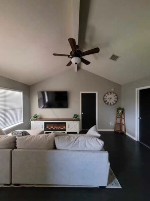 A living room with a sectional sofa, a flat-screen TV, a fireplace, a ceiling fan, and a clock on the wall. - Fantastic 3-bedroom house with a stunning patio in charming Conroe (Conroe)