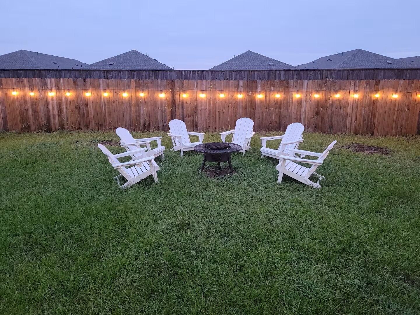 A grassy outdoor area with a wooden fence, a string of lights, and four white Adirondack chairs arranged around a small round table.