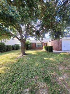 A residential property with a well-maintained lawn, a large tree providing shade, and a brick house with a garage. - Fantastic 3-bedroom house with a stunning patio in charming Conroe (Conroe)
