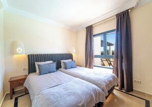 Ein Hotelzimmer mit zwei Betten, einem Fenster mit Vorhängen, Blick ins Freie und einem kleinen Tisch.