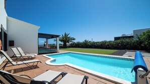 Ein Swimmingpool unter blauem Himmel, umgeben von einer Liegefläche mit Liegestühlen und Blick aufs Meer.