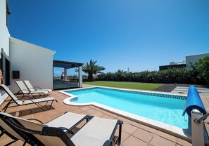 Ein Swimmingpool unter blauem Himmel, umgeben von einer Liegefläche mit Liegestühlen und Blick aufs Meer.