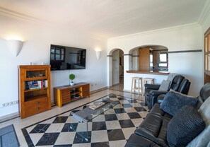 A living room with a checkered floor, a sofa, a coffee table, a bookshelf, a TV, and a kitchen area with a counter and stools. - 3 bedroom villa on a sizeable plot, close to Playa Blanca's lighthouse and the amenities of Faro Par (Playa Blanca)