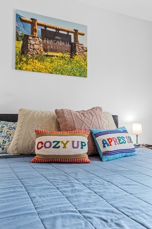 A bedroom with a blue quilted bedspread, a painting of a wooden structure with the text 'FLAGSTAFF ARIZONA 1882' above it, and two decorative pillows with the words 'COZY UP' and 'APRES SK' on them. - 2 Suite to be good! Escape to the Mountains! (Flagstaff)
