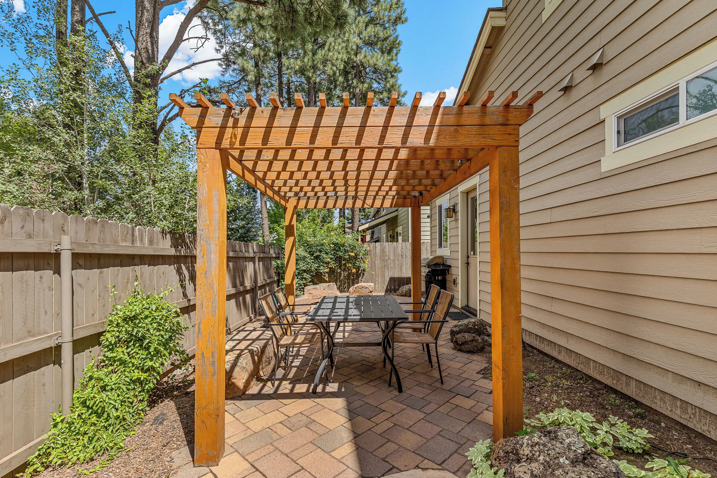 Een houten pergola met een tafel en stoelen eronder, omringd door een houten omheining en beplanting.