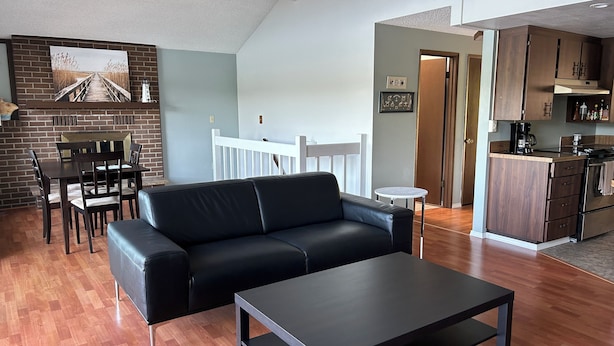 Una sala de estar moderna con un sofá de cuero negro, una mesa de centro y un comedor con pared de ladrillo y barandilla de madera.