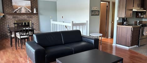 Una sala de estar moderna con un sofá de cuero negro, una mesa de centro y un comedor con pared de ladrillo y barandilla de madera.